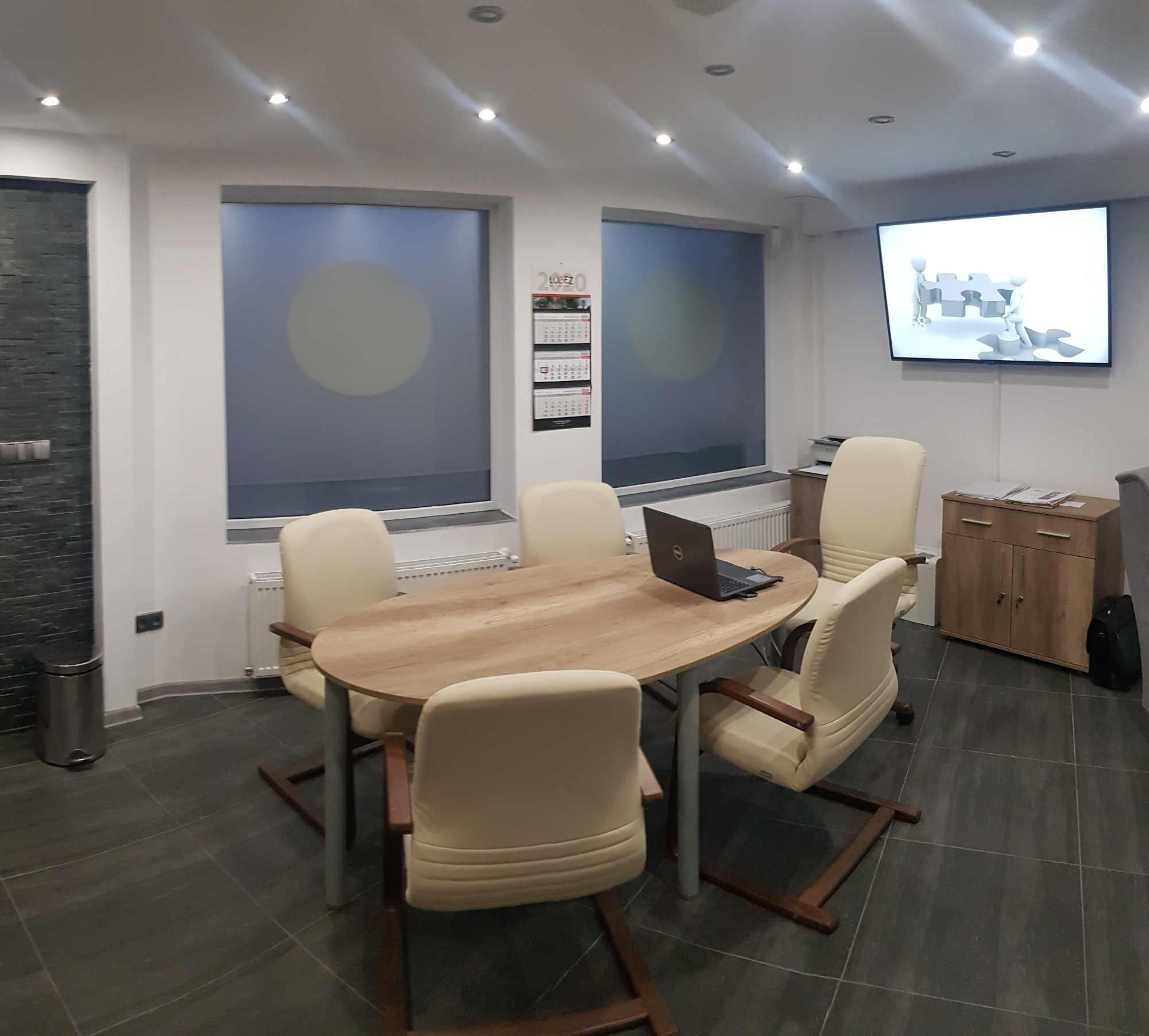 Jasne biuro z owalnym stołem, laptopem, krzesłami z drewnianymi podłokietnikami, kalendarzem na ścianie i telewizorem wyświetlającym graficzną prezentację pracy zespołowej.