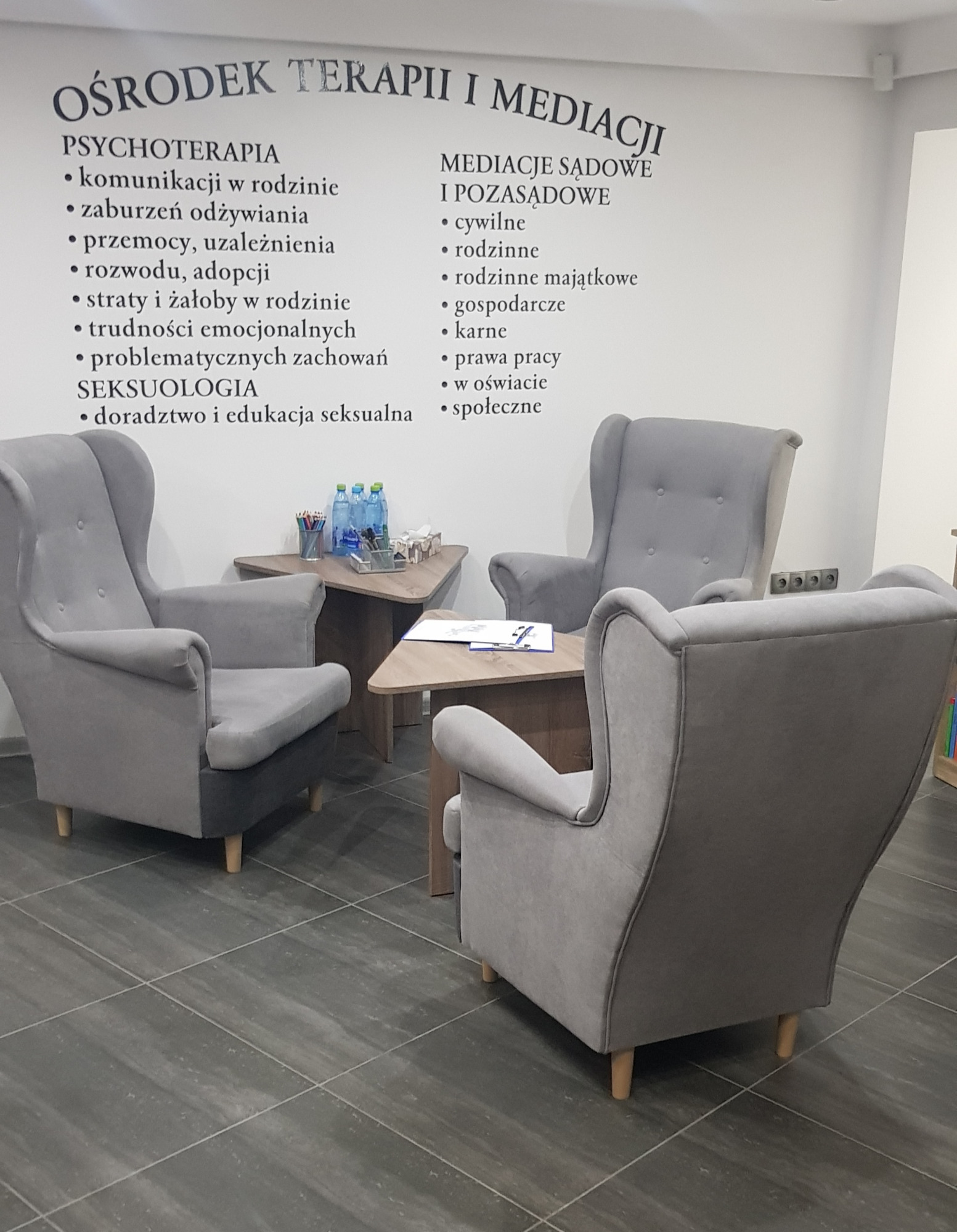 Wnętrze gabinetu terapeutycznego z trzema szarymi fotelami ustawionymi wokół małego, trójkątnego stolika, na ścianie wypisane rodzaje terapii i mediacji.