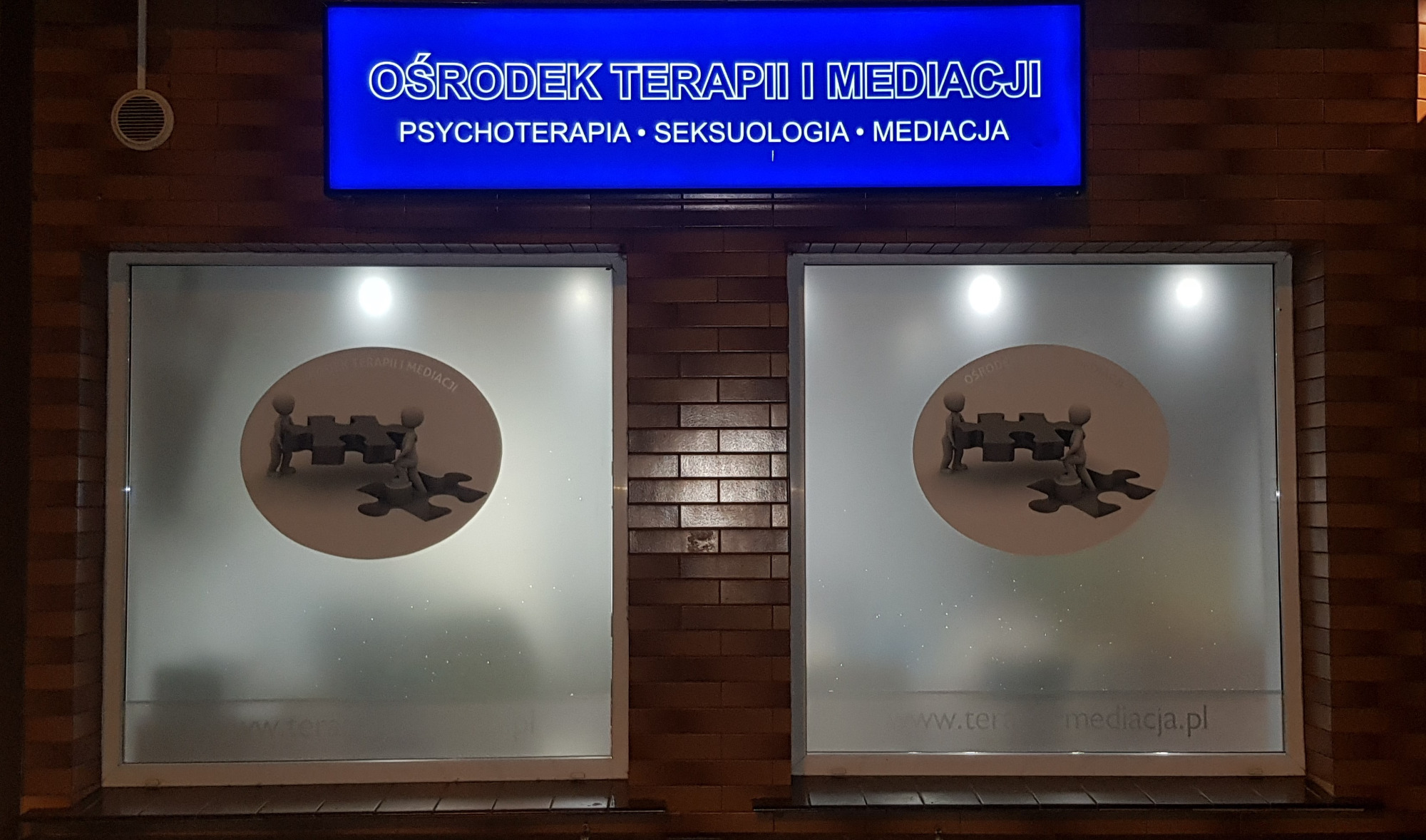 Fasada budynku Ośrodka Terapii i Mediacji z podświetlanym szyldem oraz grafikami na oknach przedstawiającymi dwie postacie układające puzzle.