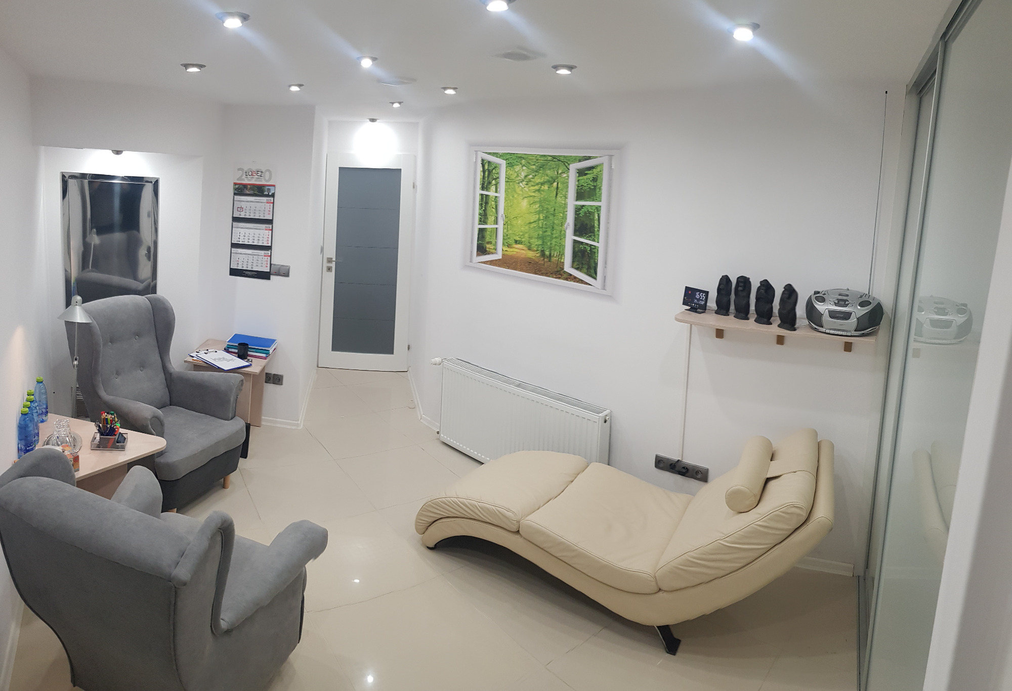 Jasne, przestronne wnętrze gabinetu z dwoma fotelami uszakami, beżową leżanką, dekoracją okienną z motywem leśnym, kalendarzem na ścianie i radiem na półce.