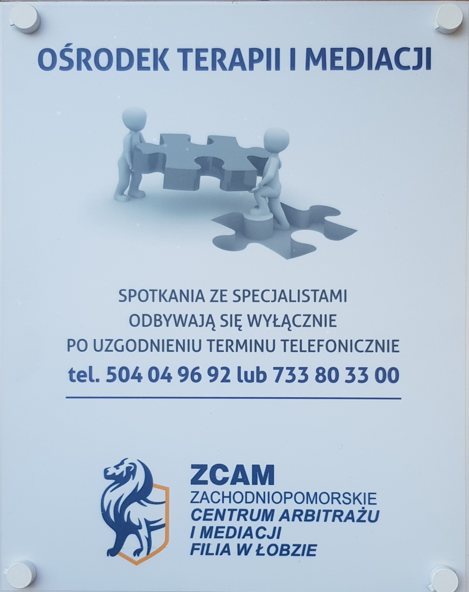 Tablica informacyjna Ośrodka Terapii i Mediacji z grafiką przedstawiającą dwie postacie układające puzzle, numerami telefonów i logo ZCAM Zachodniopomorskie Centrum Arbitrażu i Mediacji Filia...