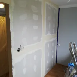 Wnętrze w trakcie remontu: ściana z płyt gipsowo-kartonowych, szpachlowane łączenia, widoczne przewody elektryczne, drabina oparta o ścianę, podłoga w panelach.