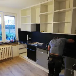 Montaż czarnego panelu ściennego w kuchni z białymi szafkami; widoczny mężczyzna w roboczych spodniach i szarej koszulce, instalujący panel nad blatem, z otwartą szafką pod spodem.