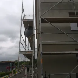 Elewacja budynku w trakcie ocieplania, widoczne rusztowania i szara, wygładzona powierzchnia ściany, fragment ogrodu z kwiatami w tle.