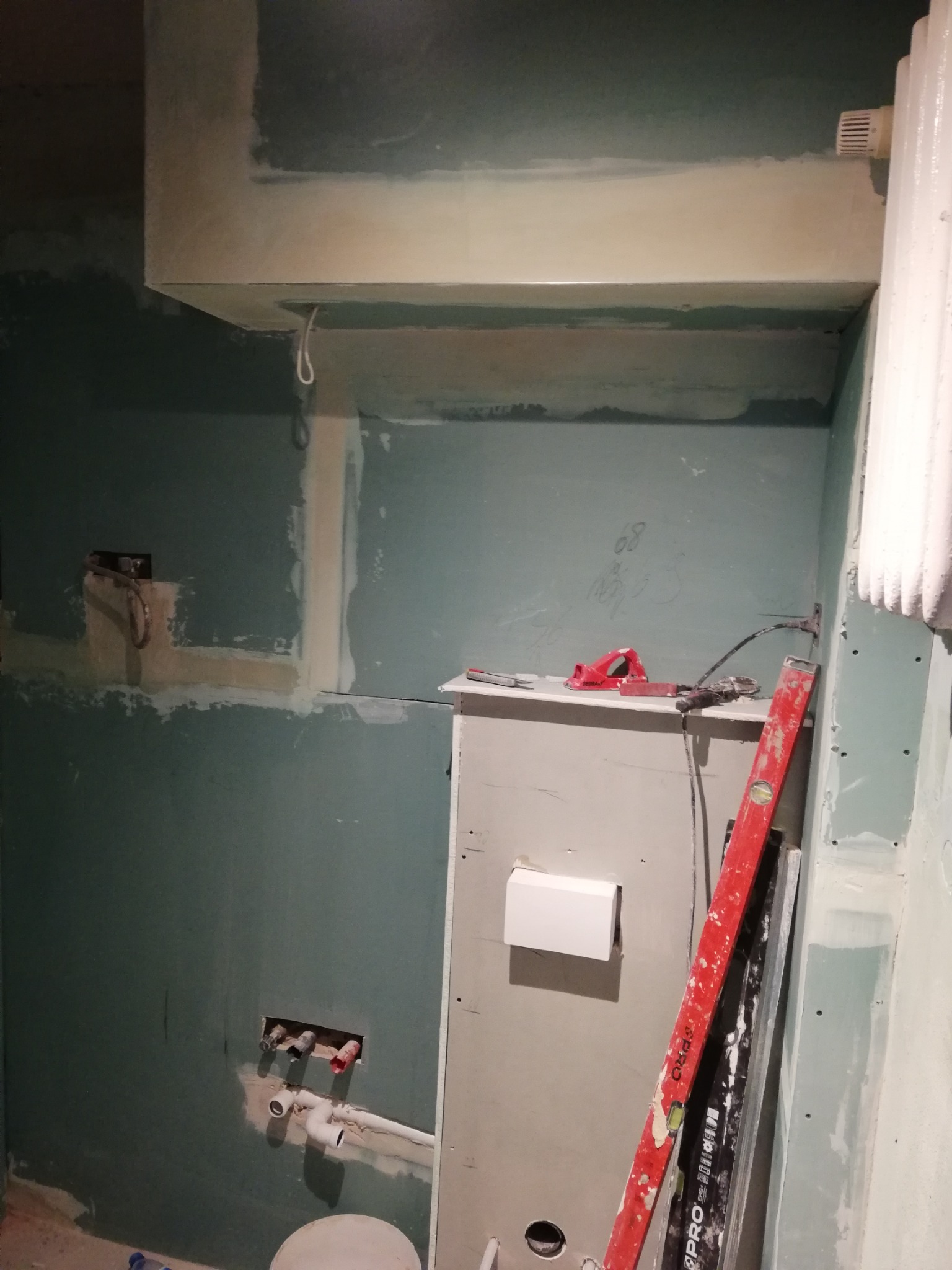 Montaż instalacji podtynkowej w łazience z widocznymi rurami, puszką elektryczną, poziomnicą i narzędziami na tle ścian wykończonych płytami gipsowo-kartonowymi przed szpachlowaniem.