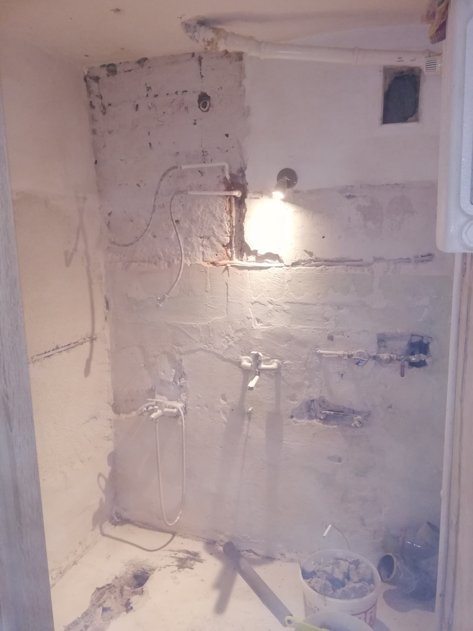 Naga ściana w łazience w trakcie remontu, z widocznymi rurami, instalacją wodną, śladami po skuciu płytek i wiadrem z gruzem na podłodze.