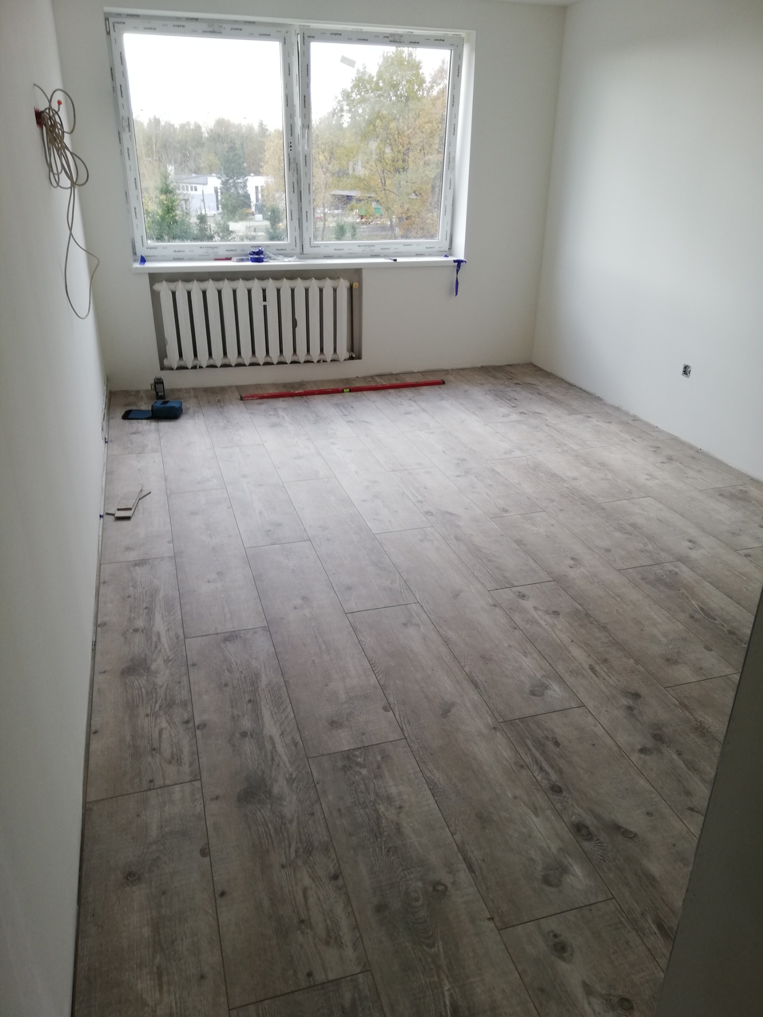 Pokój po remoncie z nową podłogą imitującą drewno, widoczne narzędzia pomiarowe i grzejnik pod oknem.
