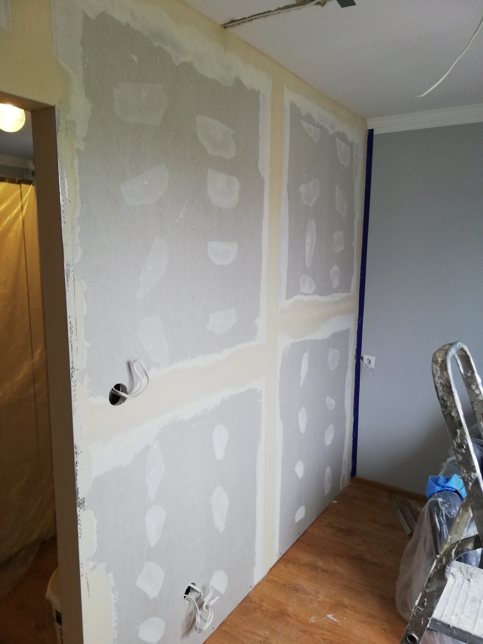 Wnętrze w trakcie remontu: ściana z płyt gipsowo-kartonowych, szpachlowane łączenia, widoczne przewody elektryczne, drabina oparta o ścianę, podłoga w panelach.