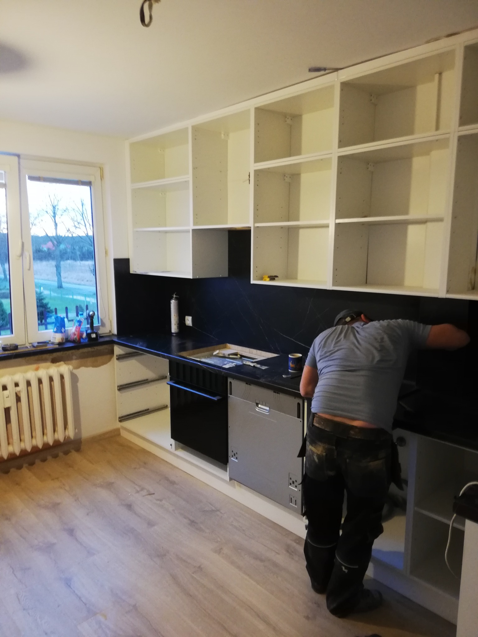Montaż czarnego panelu ściennego w kuchni z białymi szafkami; widoczny mężczyzna w roboczych spodniach i szarej koszulce, instalujący panel nad blatem, z otwartą szafką pod spodem.