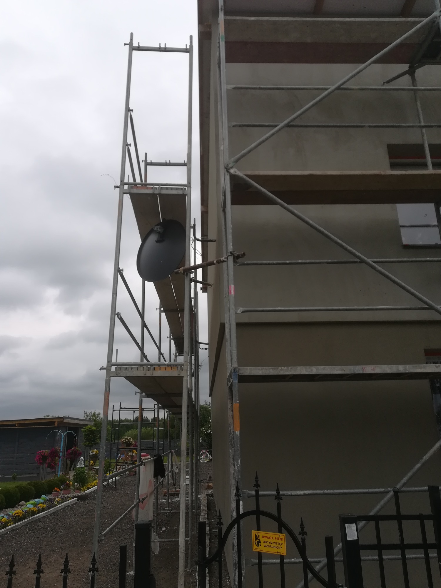 Elewacja budynku w trakcie ocieplania, widoczne rusztowania i szara, wygładzona powierzchnia ściany, fragment ogrodu z kwiatami w tle.