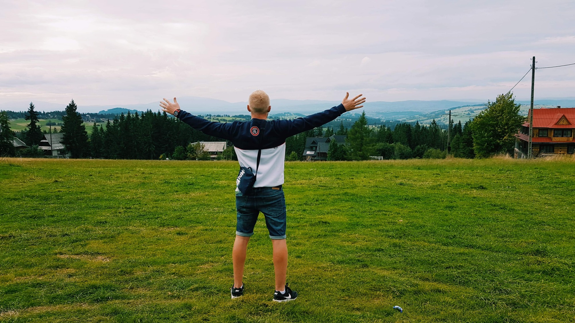 Mężczyzna stojący na zielonym wzgórzu z rozłożonymi ramionami, w tle widok na góry i domy z czerwonymi dachami.