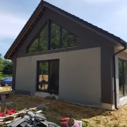 Nowoczesny dom jednorodzinny z szarą elewacją i dużymi oknami trójkątnymi, widoczne ślady prac wykończeniowych, resztki dachówek i puste wiadra po farbie na trawniku.