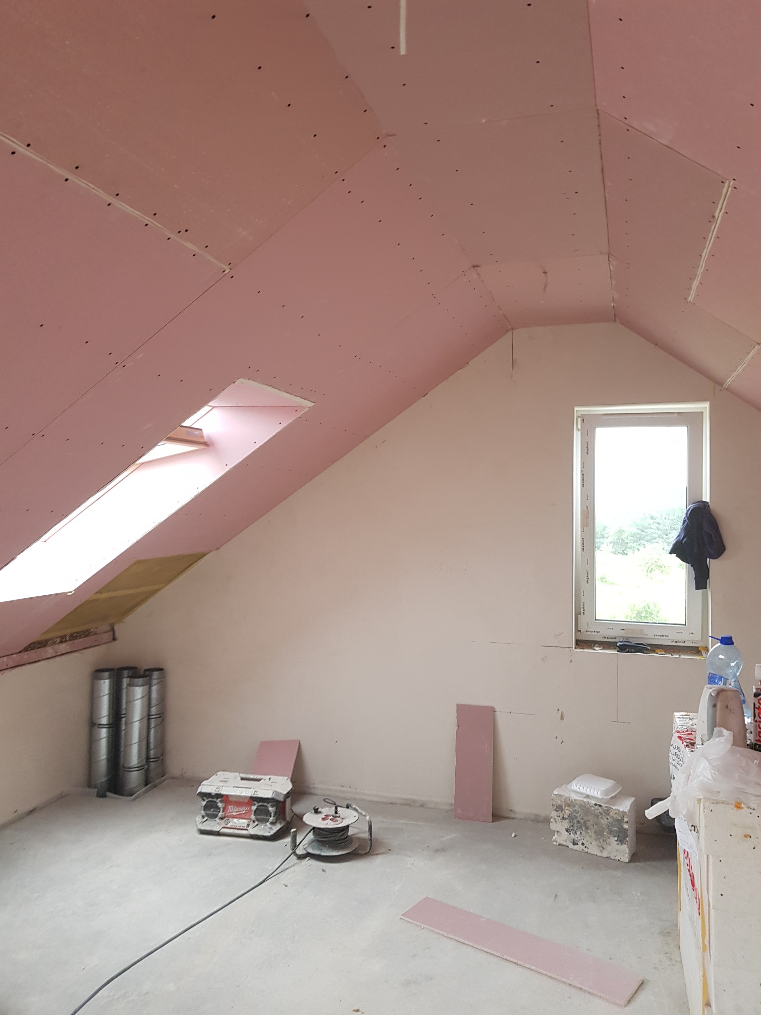 Poddasze w trakcie prac wykończeniowych: zamontowane płyty kartonowo-gipsowe w kolorze różowym, okno dachowe, okno pionowe z widokiem na zieleń, narzędzia i materiały budowlane rozłożone na podłodze.