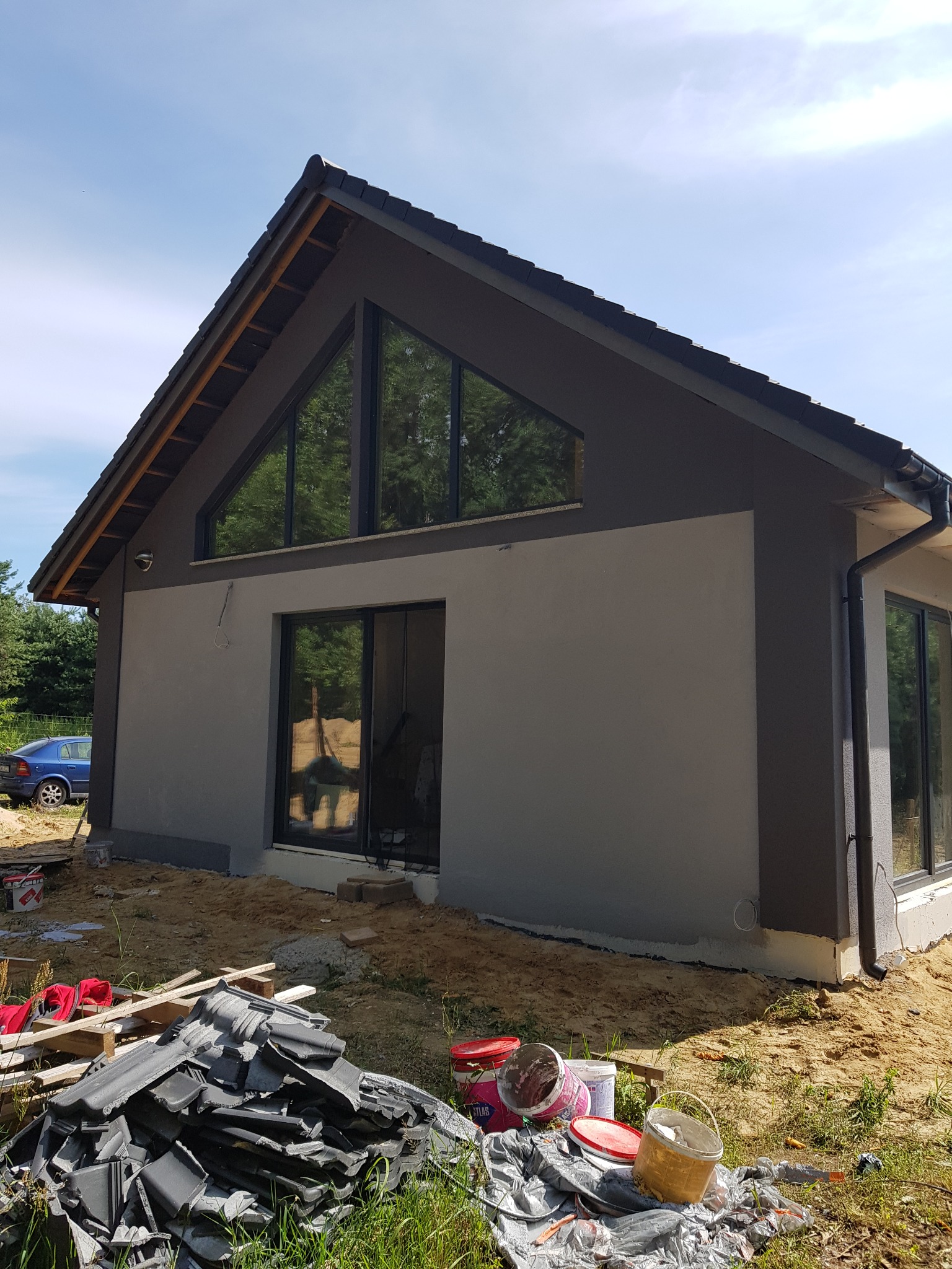 Nowoczesny dom jednorodzinny z szarą elewacją i dużymi oknami trójkątnymi, widoczne ślady prac wykończeniowych, resztki dachówek i puste wiadra po farbie na trawniku.