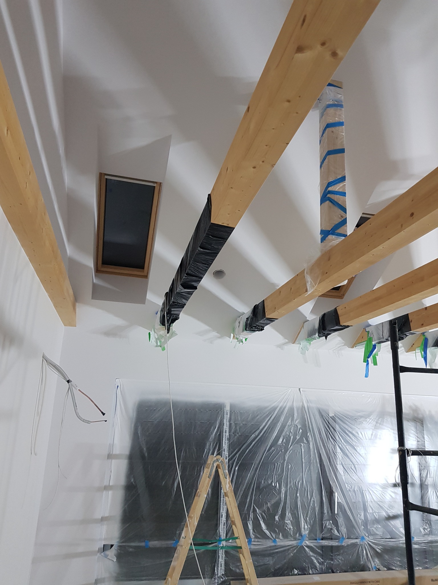Poddasze w trakcie prac wykończeniowych: drewniane belki stropowe, okno dachowe, zabezpieczona folią ściana, drabina i rusztowanie.