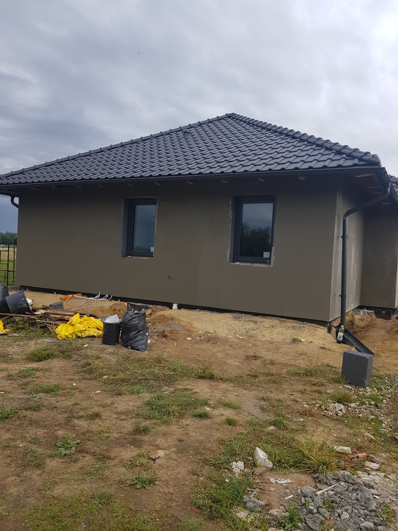 Dom jednorodzinny z szarą elewacją i czarną dachówką, widoczne worki i materiały budowlane wokół, teren w trakcie prac wykończeniowych.