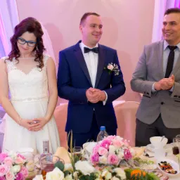 Panna młoda w białej sukni, pan młody w granatowym smokingu i gość w szarej marynarce stoją za stołem z kwiatami i napojami podczas weselnego przyjęcia.