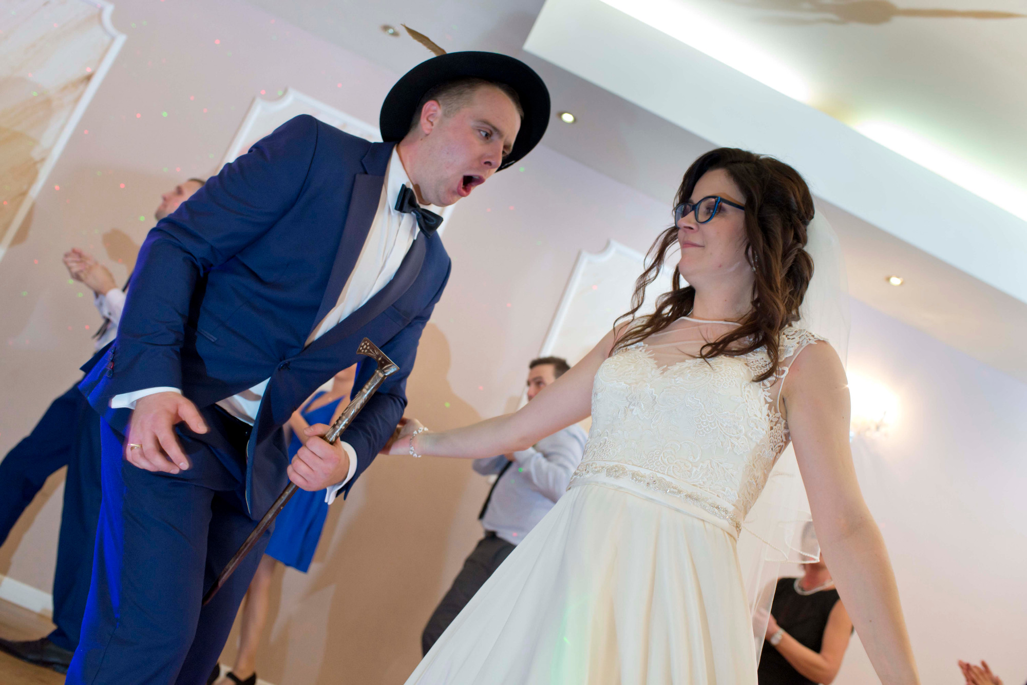 Rozbawiony pan młody w kapeluszu i fraku z laską prowadzi taniec z uśmiechniętą panną młodą w białej sukni podczas wesela, w tle inni goście.