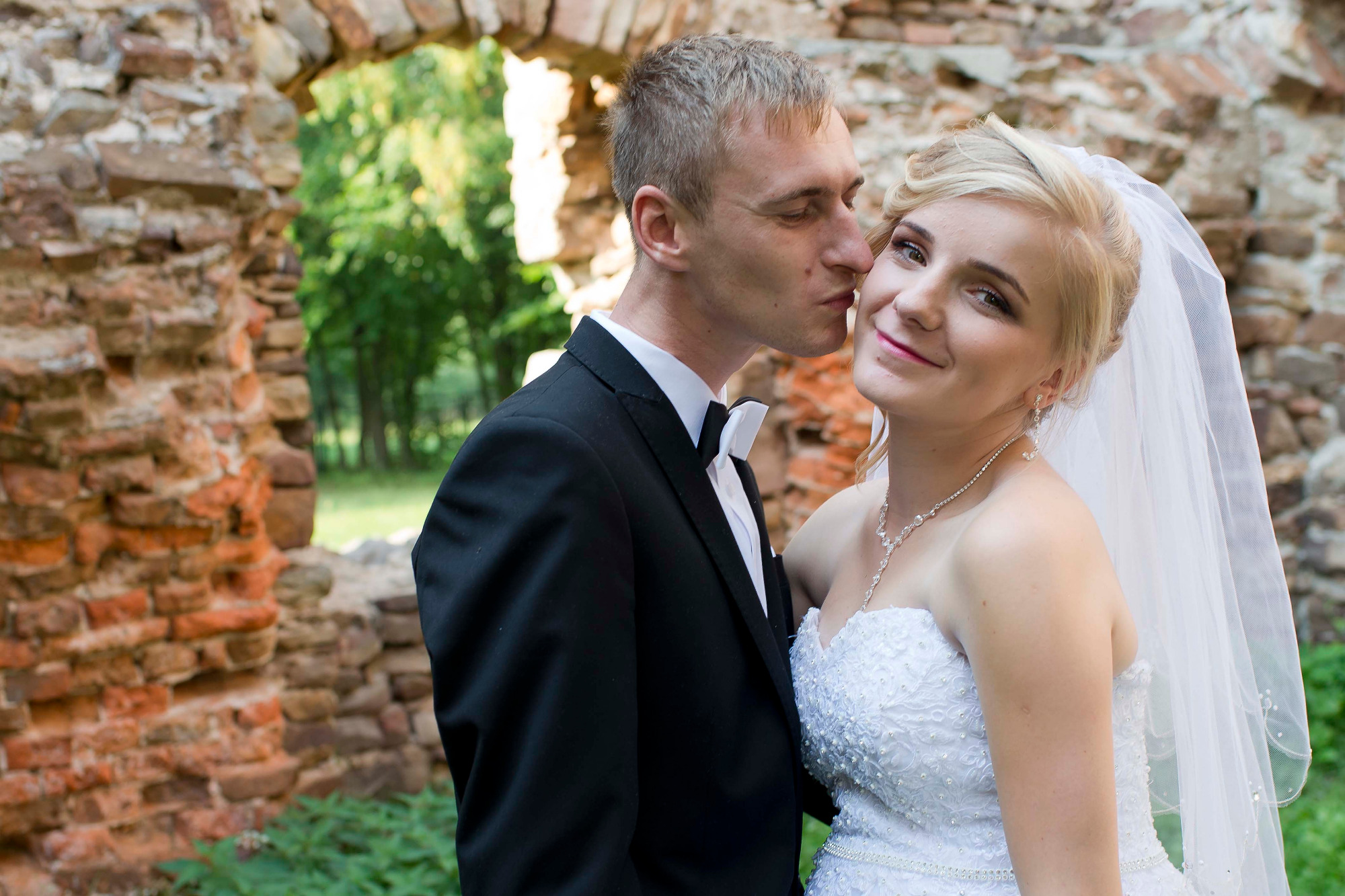 Panna młoda w białej sukni z welonem, uśmiechnięta, obejmowana przez pana młodego w czarnym garniturze z białą muchą, na tle kamiennych ruin z zielenią w tle.