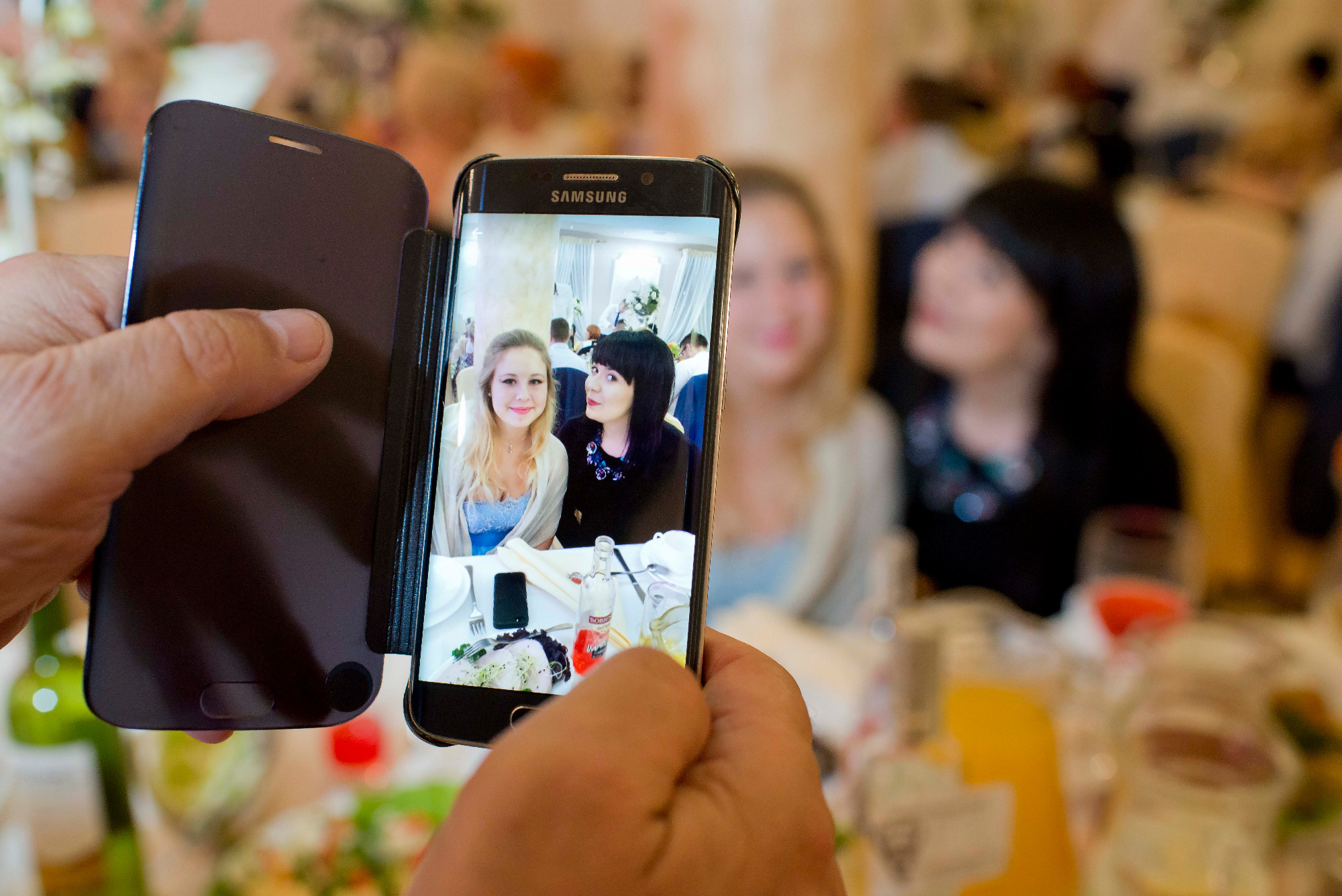 Osoba fotografuje telefonem Samsung zdjęcie dwóch kobiet siedzących przy stole podczas wydarzenia, w tle rozmazani goście.
