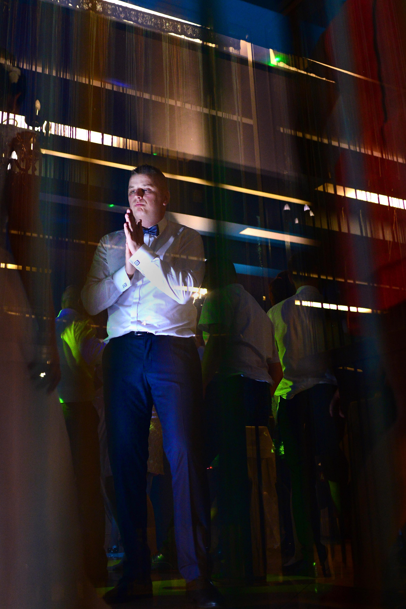 Mężczyzna w białej koszuli i muszce, stojący na tle dekoracji świetlnej i innych osób, w trakcie wydarzenia.
