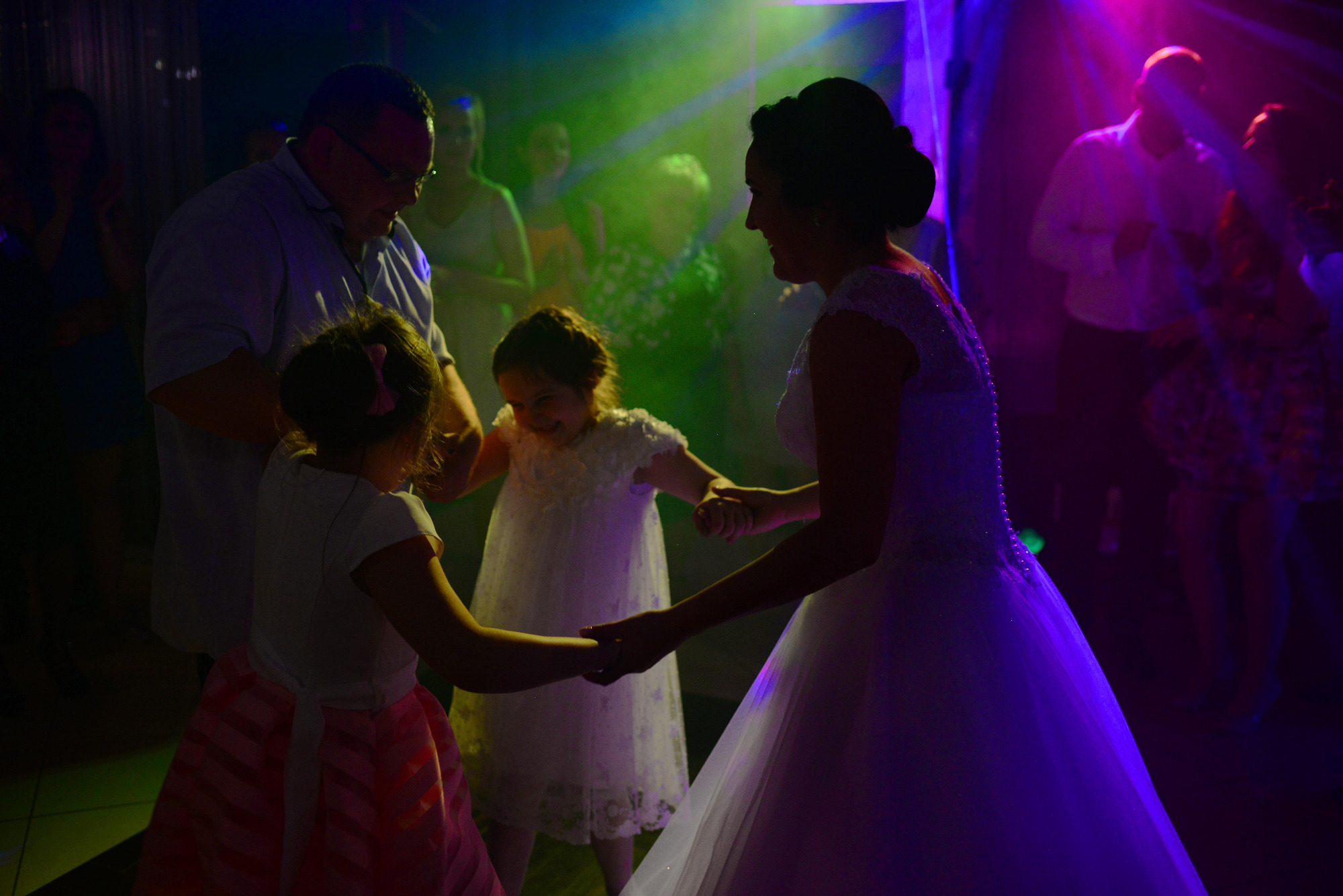 Panna młoda w białej sukni trzyma za ręce dwie dziewczynki w białych i różowych sukienkach, tańczące na parkiecie z kolorowym oświetleniem, w tle goście.