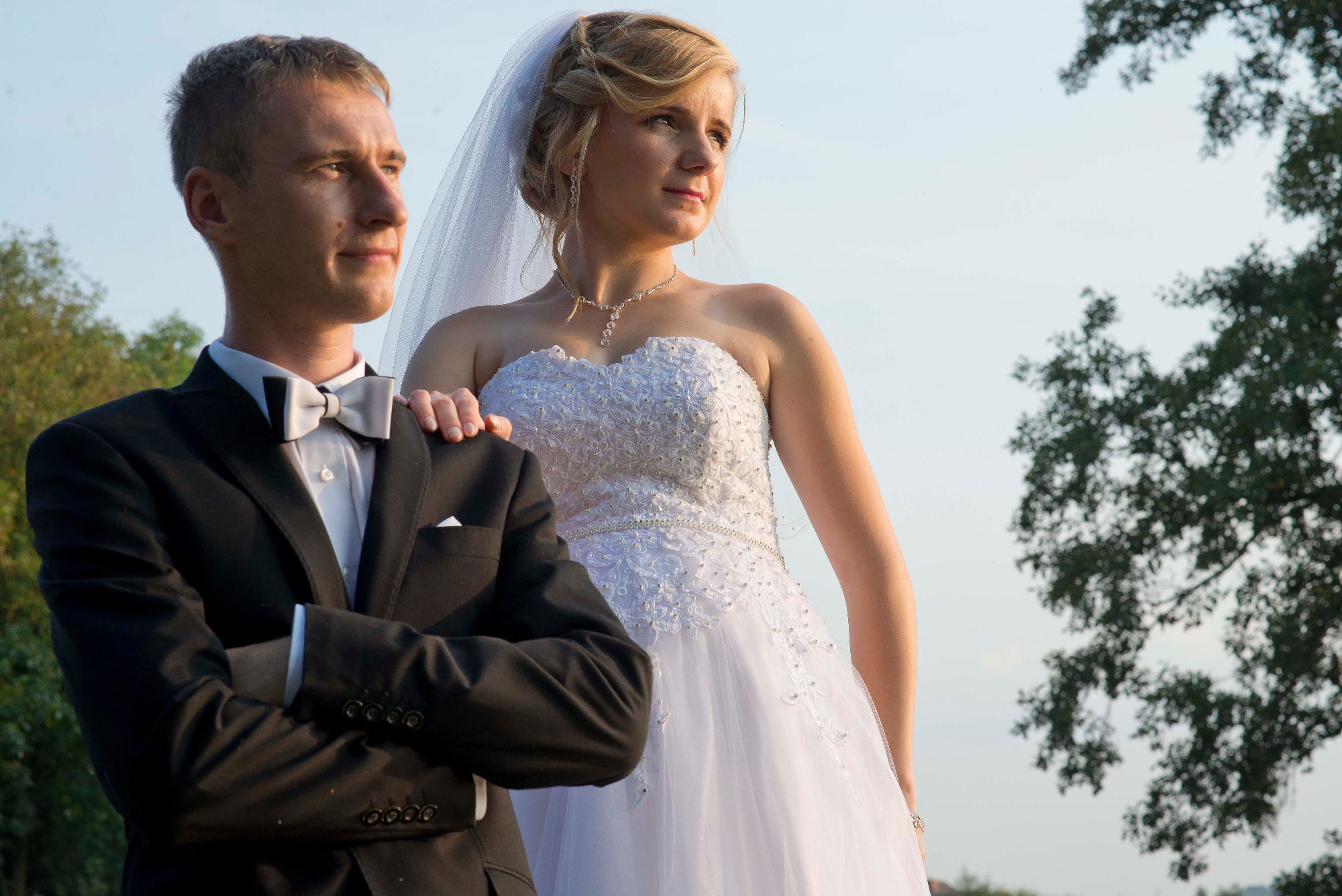 Para młoda: panna młoda w białej sukni z welonem i pan młody w czarnym smokingu z muszką, stojący na tle drzew i jasnego nieba.