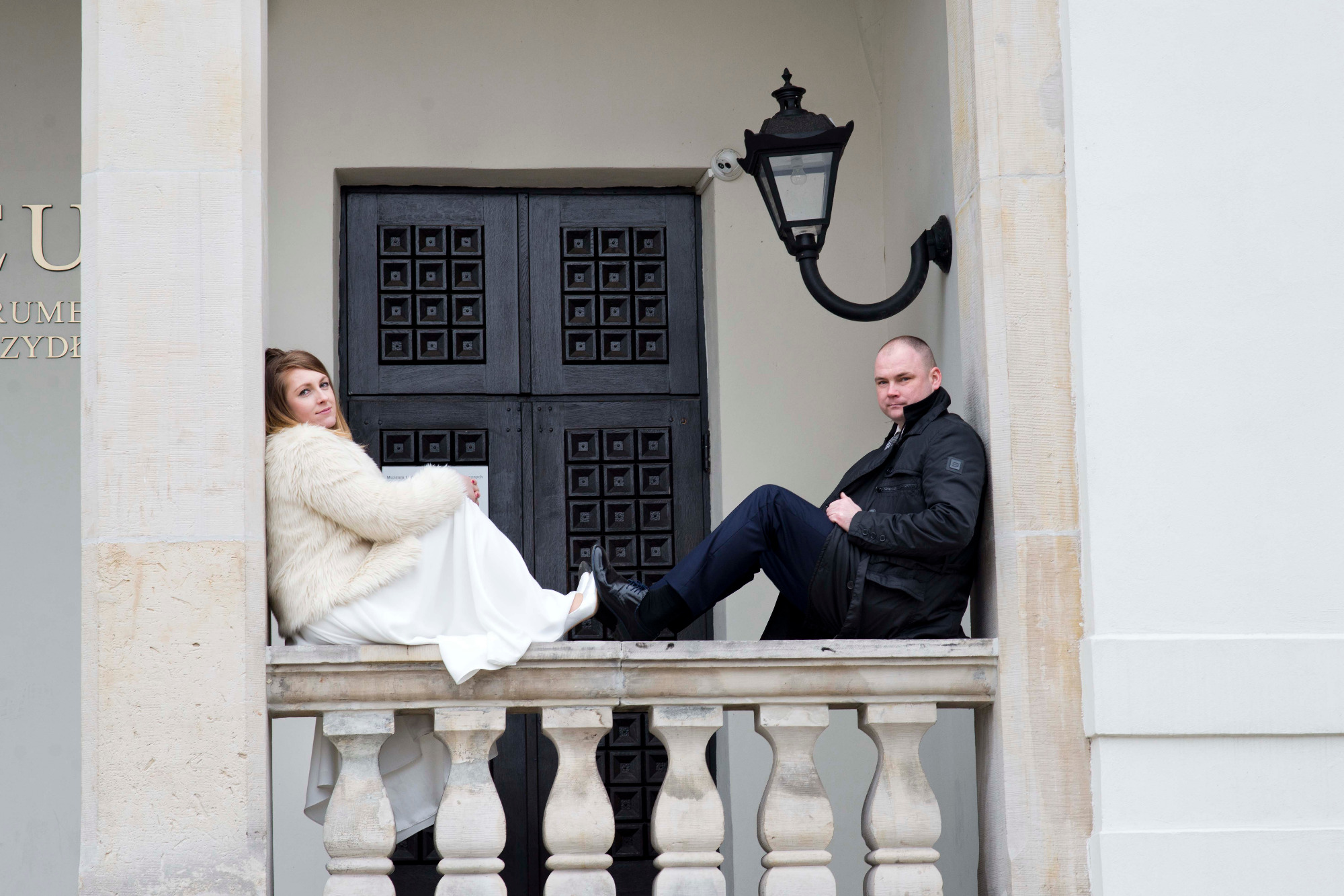 Para siedząca na balkonie z kamienną balustradą, ona w białej sukni i futrze, on w czarnej kurtce, w tle czarne drzwi i lampa.