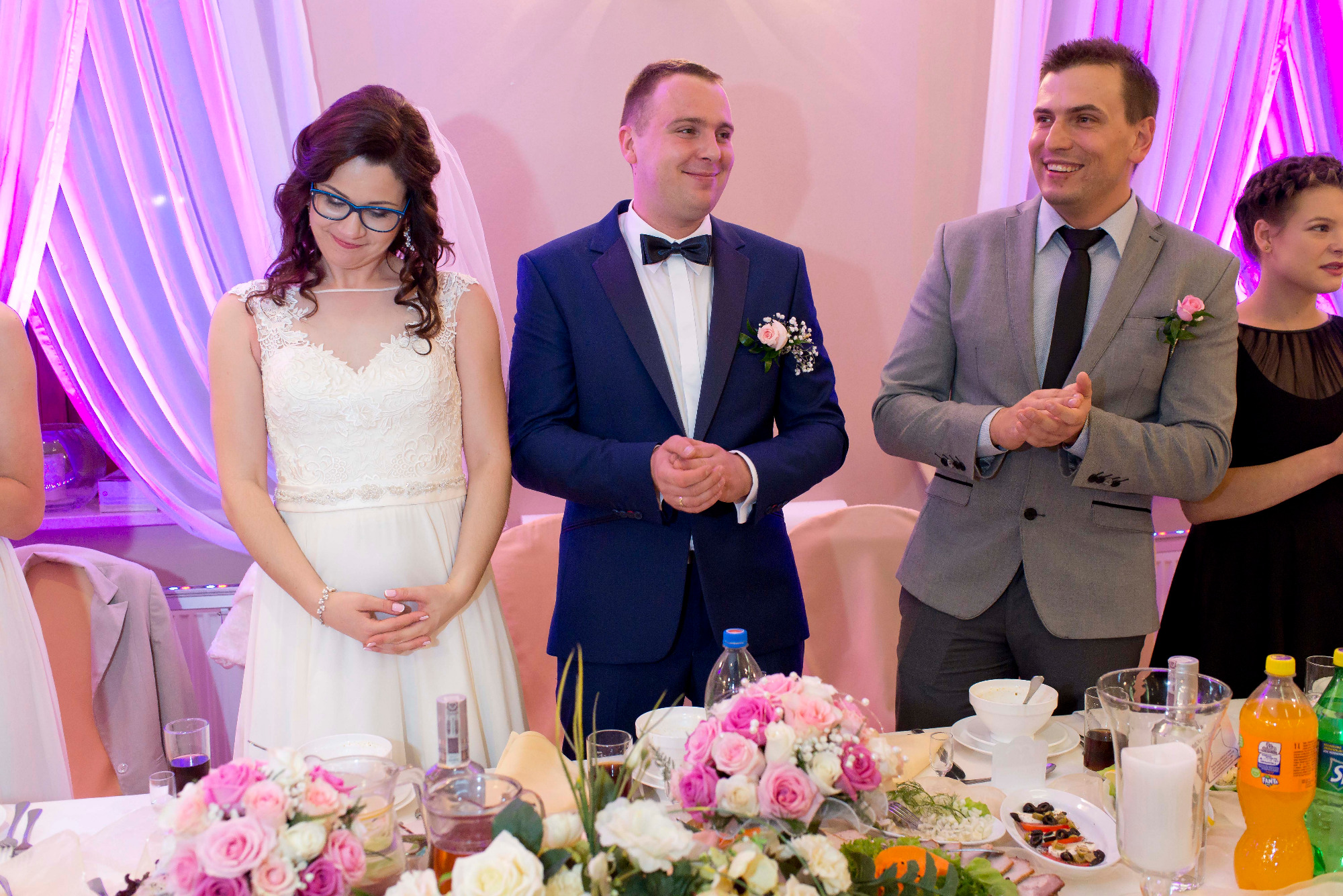 Panna młoda w białej sukni, pan młody w granatowym smokingu i gość w szarej marynarce stoją za stołem z kwiatami i napojami podczas weselnego przyjęcia.