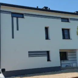 Nowoczesny dom jednorodzinny z jasną elewacją i grafitowymi akcentami dekoracyjnymi, widok z perspektywy ulicy, z przodu zaparkowany biały samochód dostawczy.