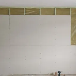 Ściana w trakcie montażu płyt gipsowo-kartonowych na stelażu z widoczną izolacją z wełny mineralnej, częściowo zakrytej folią, z drewnianą drabiną opartą o ścianę po lewej stronie i wkrętarką...