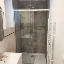 Nowoczesna kabina prysznicowa z deszczownicą i szklanymi drzwiami przesuwnymi, umywalka z baterią w odcieniach srebra oraz grzejnik drabinkowy w łazience po remoncie w Kołaczkowicach.