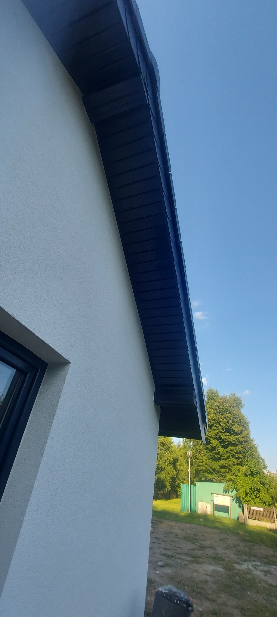 Fragment ocieplonej elewacji budynku w Kołaczkowicach z widoczną podbitką dachową i oknem. Ściana w jasnym odcieniu, podbitka ciemna, na zewnątrz zieleń drzew i błękit nieba.