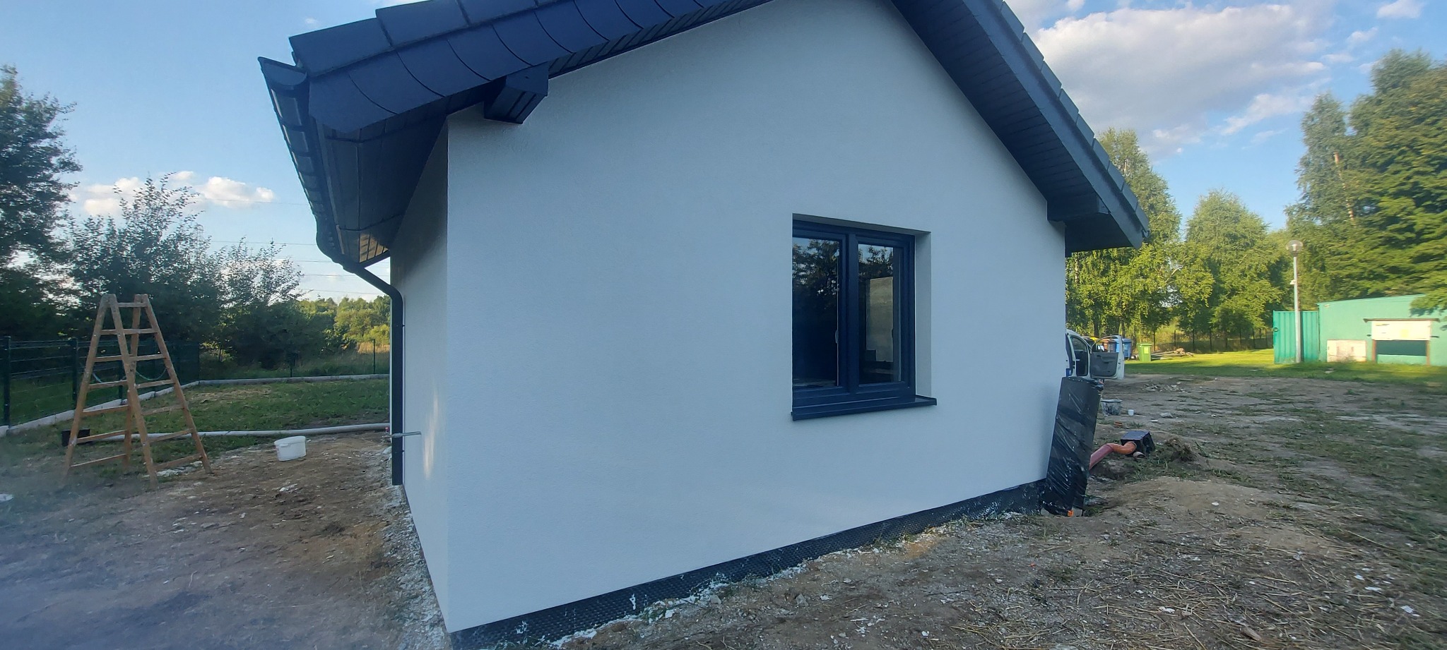 Świeżo otynkowana ściana domu jednorodzinnego w jasnym kolorze, z widocznym oknem w ciemnej ramie, czarnym dachem i fragmentem rynny, na tle zieleni drzew i nieba.