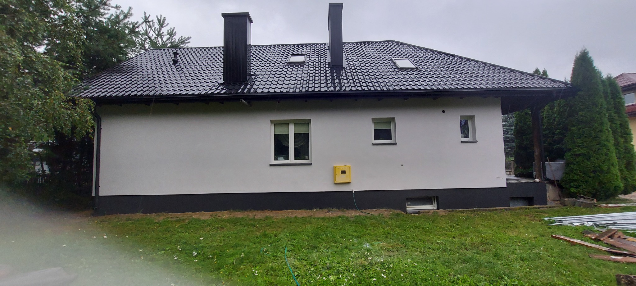 Świeżo pomalowana elewacja domu jednorodzinnego z czarnym dachem i kominami, widoczna skrzynka gazowa na ścianie, trawnik z elementami budowlanymi w tle.