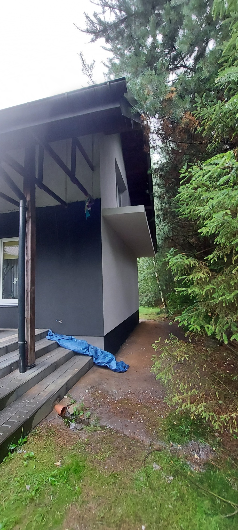 Fragment ocieplonego budynku z szarą elewacją, przykryty niebieską folią ochronną na schodach, z widocznym fragmentem ogrodu i drzew