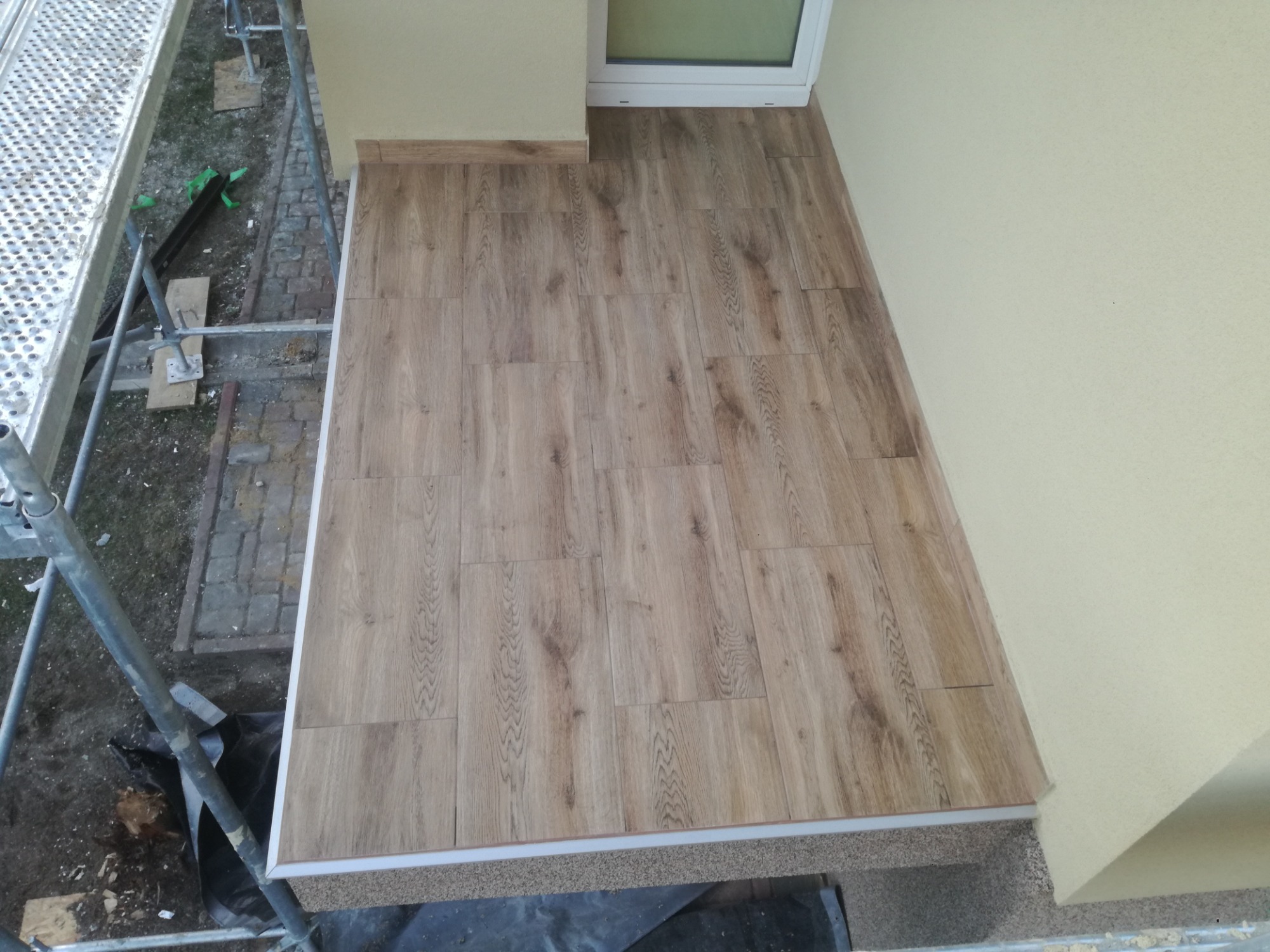 Ukończona podłoga balkonu wyłożona panelami imitującymi drewno, widoczne rusztowanie i fragment elewacji budynku.