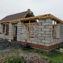 Krakusserwis - Budowa domu jednorodzinnego w trakcie: mury z bloczków betonowych na fundamencie z cegły, drewniane nadproża okienne, częściowo ułożony dach z brązowej blachodachówki, widoczny stos gruzu i opon...