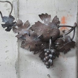Krakusserwis - Kuta, metalowa dekoracja ścienna przedstawiająca winogrona z liśćmi, przymocowana do szarego, betonowego filara z pomarańczowymi śladami farby.