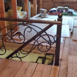 Krakusserwis - Kuta balustrada schodowa z motywami roślinnymi w trakcie montażu na drewnianym poddaszu, widoczne materiały budowlane i narzędzia.