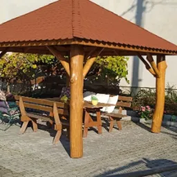 Drewniana altana z czerwoną dach&oacute;wką, wyposażona w st&oacute;ł i ławki, otoczona zielenią i brukowaną nawierzchnią.