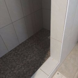 Krakusserwis - Wykończony brodzik z ciemną mozaiką na podłodze i jasnymi płytkami na ścianach, widoczne białe listwy wykończeniowe, ujęcie z góry pod kątem.