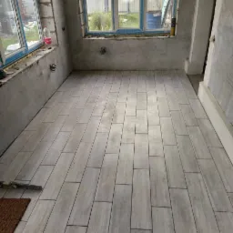 Ułożone podłużne płytki podłogowe imitujące drewno w surowym wnętrzu z oknami, widoczne narzędzia i butelka na parapecie.