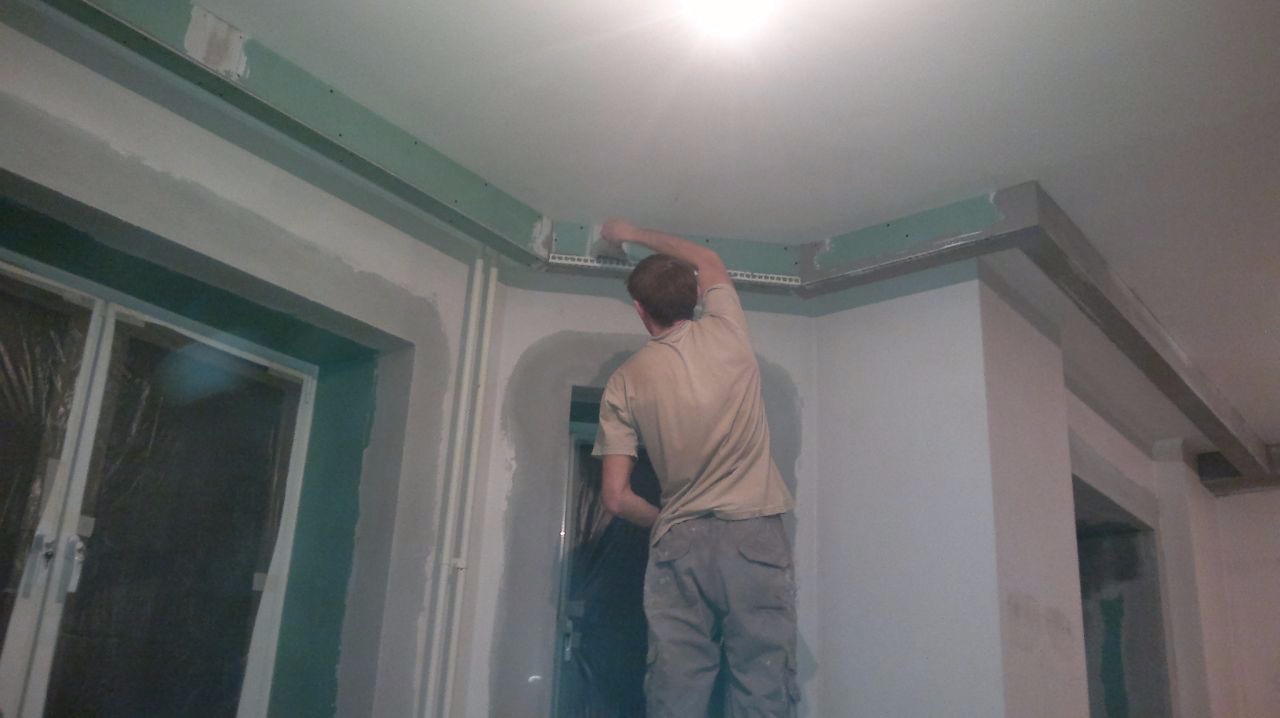 Pracownik na drabinie szpachluje łączenie płyty na suficie podwieszanym, widoczne metalowe profile i siatka narożna.