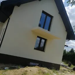 Nowoczesny dom jednorodzinny z jasną elewacją i ciemnym dachem, widoczne dwa okna z ciemnymi ramami oraz wystający element architektoniczny nad oknem, teren wokół domu w trakcie prac wykończeniowych.