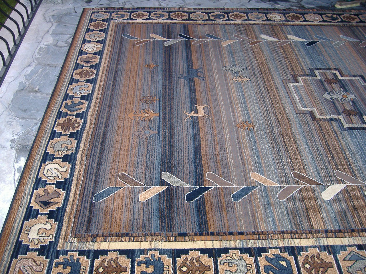 Wielobarwny dywan z motywami zwierzęcymi i geometrycznymi, leżący na szarym betonowym podłożu, widoczny z perspektywy lekko z góry.