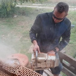 Fenstahab - Pracownik w okularach ochronnych przecina bloczki ceramiczne piłą spalinową na zewnątrz, unoszący się pył.