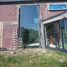 Montaż dużego okna panoramicznego w surowej bryle domu z czerwonej cegły, widoczne drabiny i narzędzia, trawa na pierwszym planie.