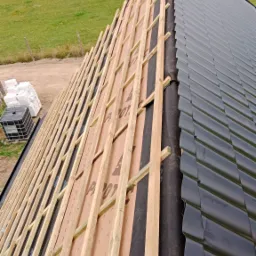 Montaż czarnej blachodachówki na drewnianej konstrukcji dachu, widok z góry na częściowo pokryty dach i zielone łąki w tle. Prace dekarskie w toku.