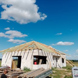 Fenstahab - Niedokończony dom z białych bloczków pod błękitnym niebem z drewnianą konstrukcją dachu, widoczne materiały budowlane i przyczepa.